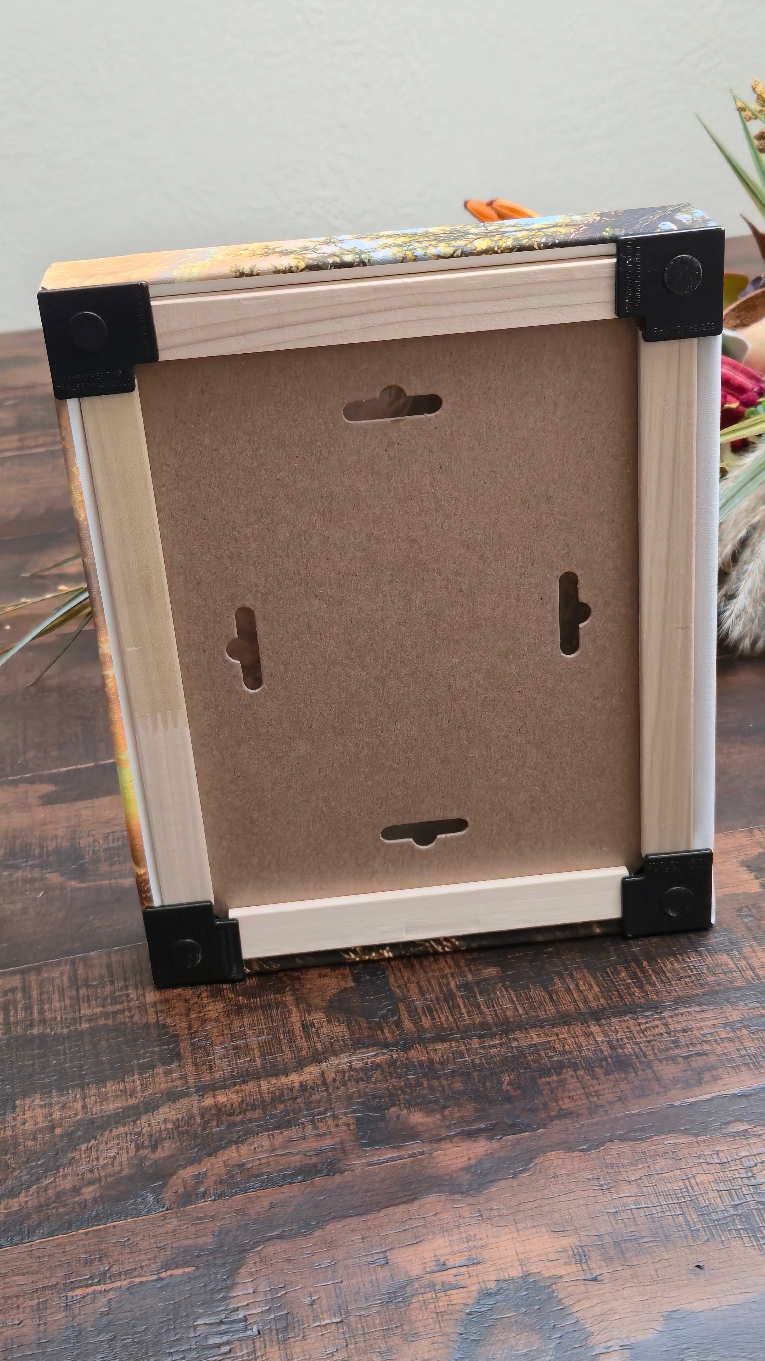 The back view of a canvas picture frame with black corner protectors, resting on a dark wood-grained surface.